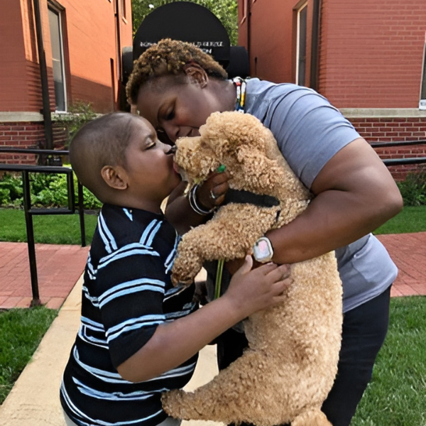 A young boy kisses a tan Doodle dog that a woman is holding up to his face. 