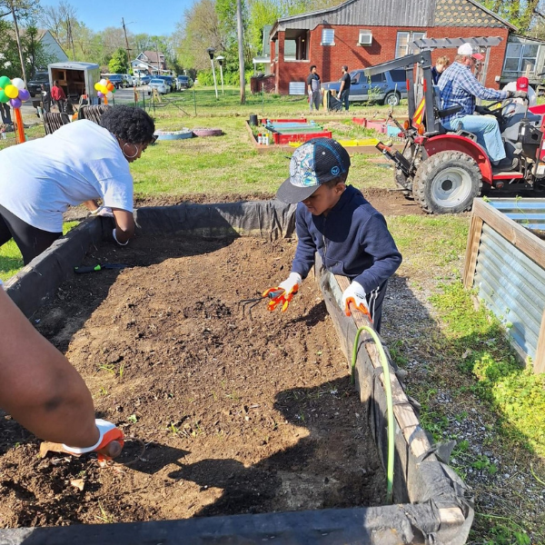People work together to prepare dirt in a garden bed for planting. A man driving a tractor is in the background.